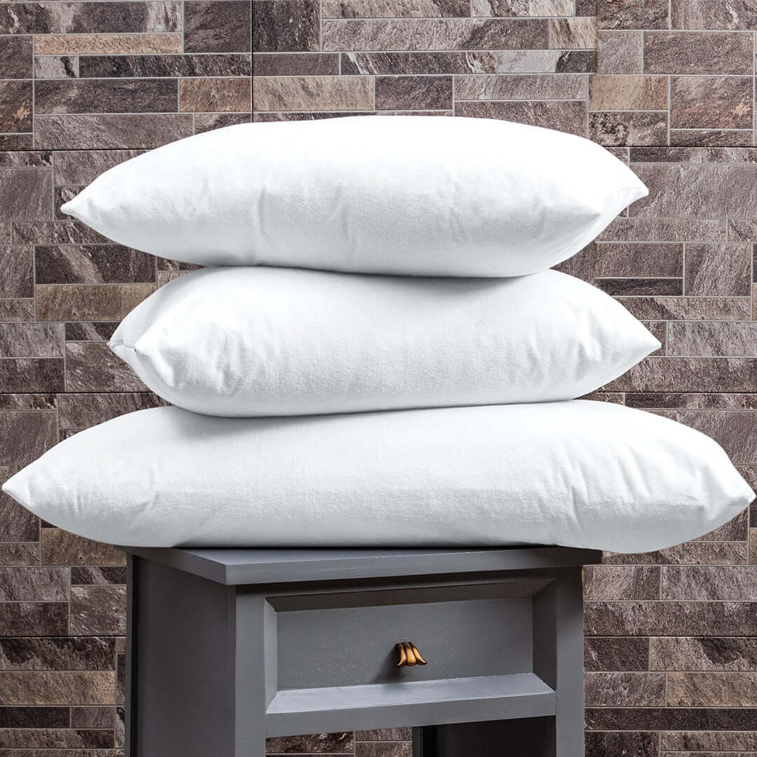 Three white pillows are stacked on top of each other on a small gray nightstand, which has a drawer and books on the shelf, with a brick-patterned wall in the background.