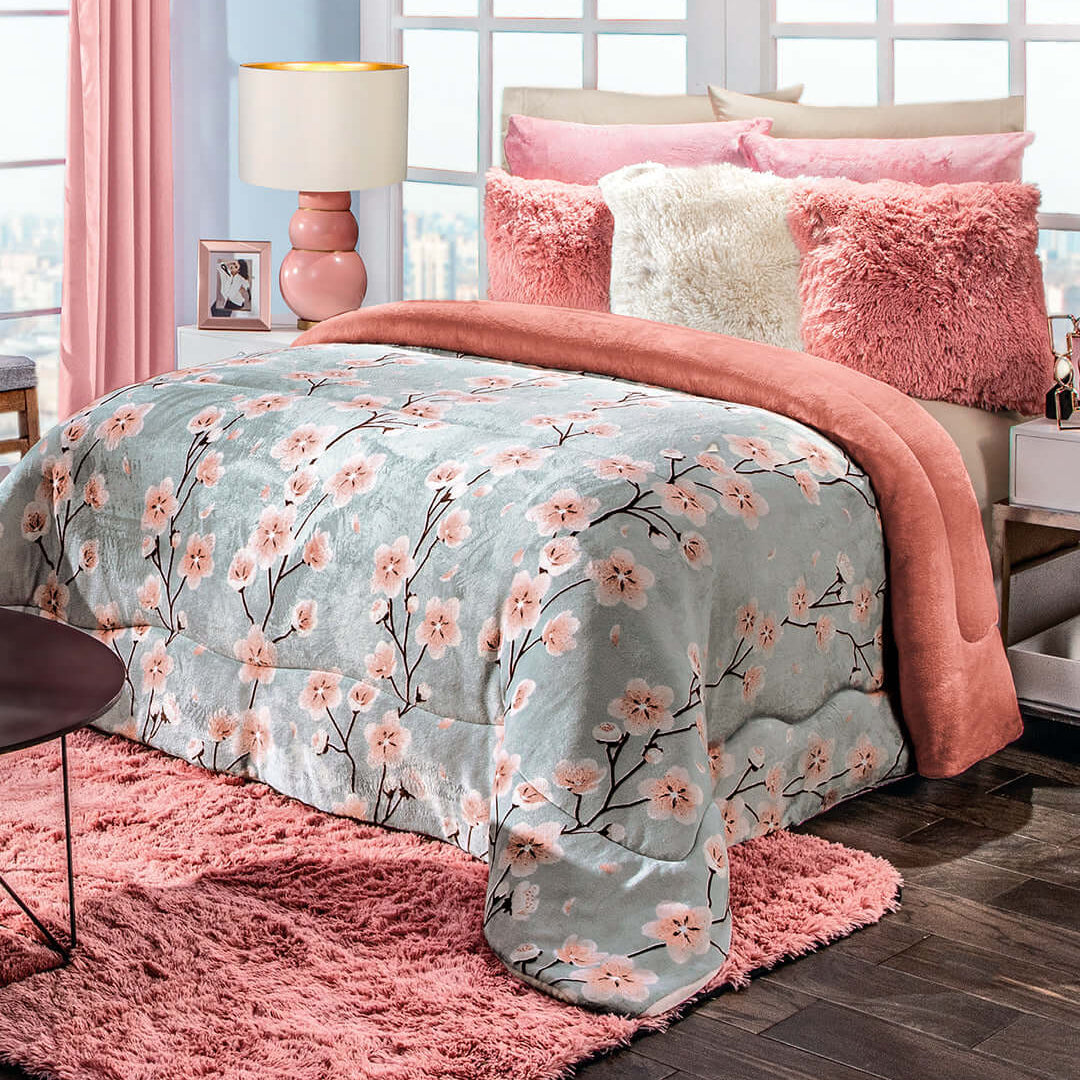 A cozy bedroom with a floral comforter, pink throw blanket, fluffy pillows, pink rug, and pink lamp. Light blue walls and large windows create a bright and inviting atmosphere.