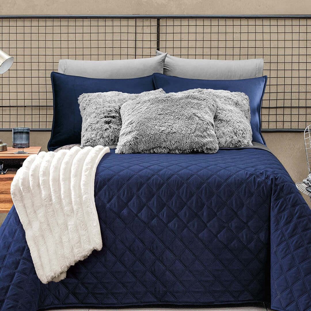 A neatly made bed with a navy blue quilt, two gray fluffy pillows, two navy pillows, and two large light gray pillows, topped with a folded white blanket at the foot, against a beige wall.