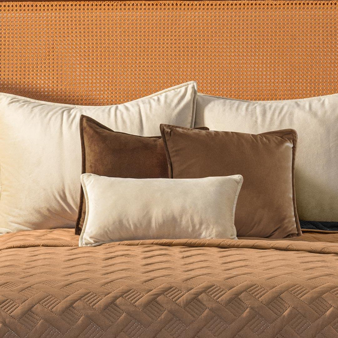A neatly made bed with a tan textured blanket, several cream and brown velvet pillows, and a rattan headboard against a light wall.