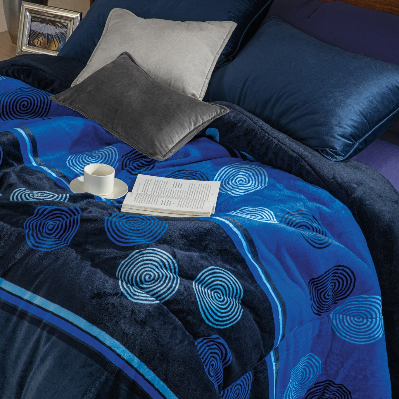 A cozy bed with blue and grey pillows, a blue-patterned blanket, an open book, and a cup on top of the blanket. A wooden headboard and nightstand with a lamp are in the background.