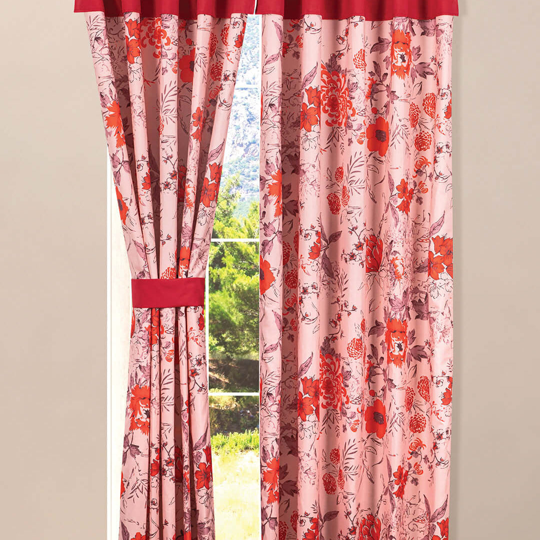 A window with pink and red floral curtains; one side is tied back with a matching band, while the other side hangs straight. Sunlight and greenery are visible outside through the window.