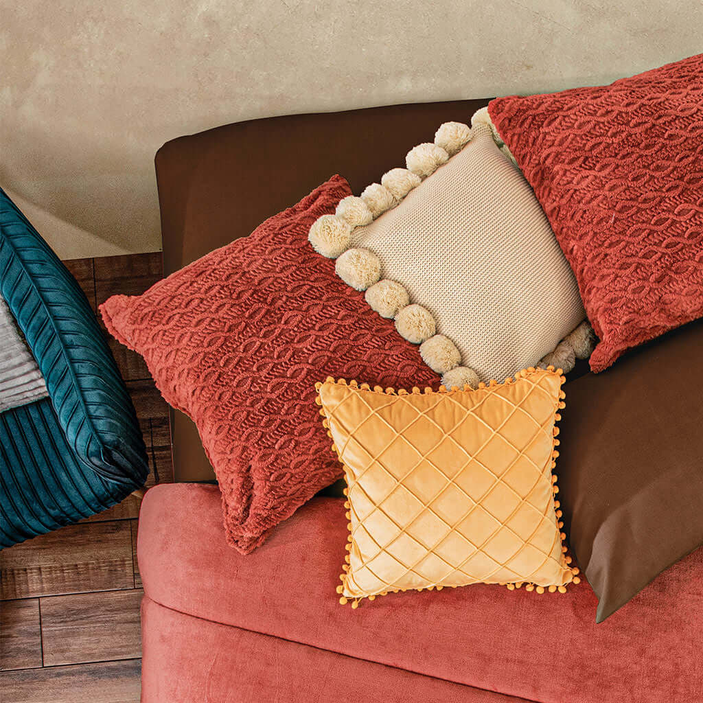 A cozy arrangement of textured pillows in red, cream, and yellow with pom-pom and quilted details, placed on a brown and red upholstered sofa near a blue corduroy seat.