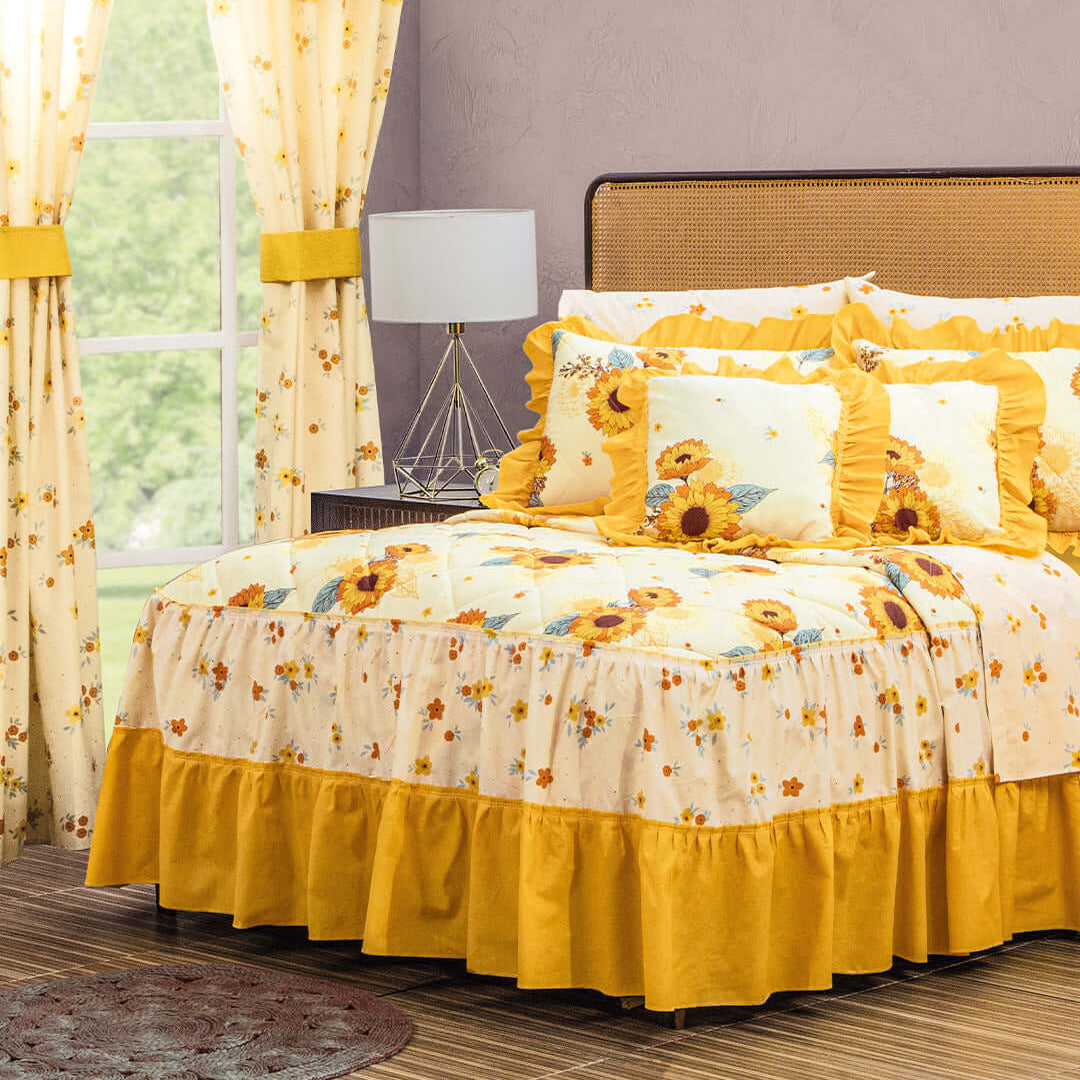 A bedroom with a bed dressed in yellow and sunflower-patterned bedding, matching sunflower curtains, decorative pillows, a bedside lamp, and a window showing greenery outside.