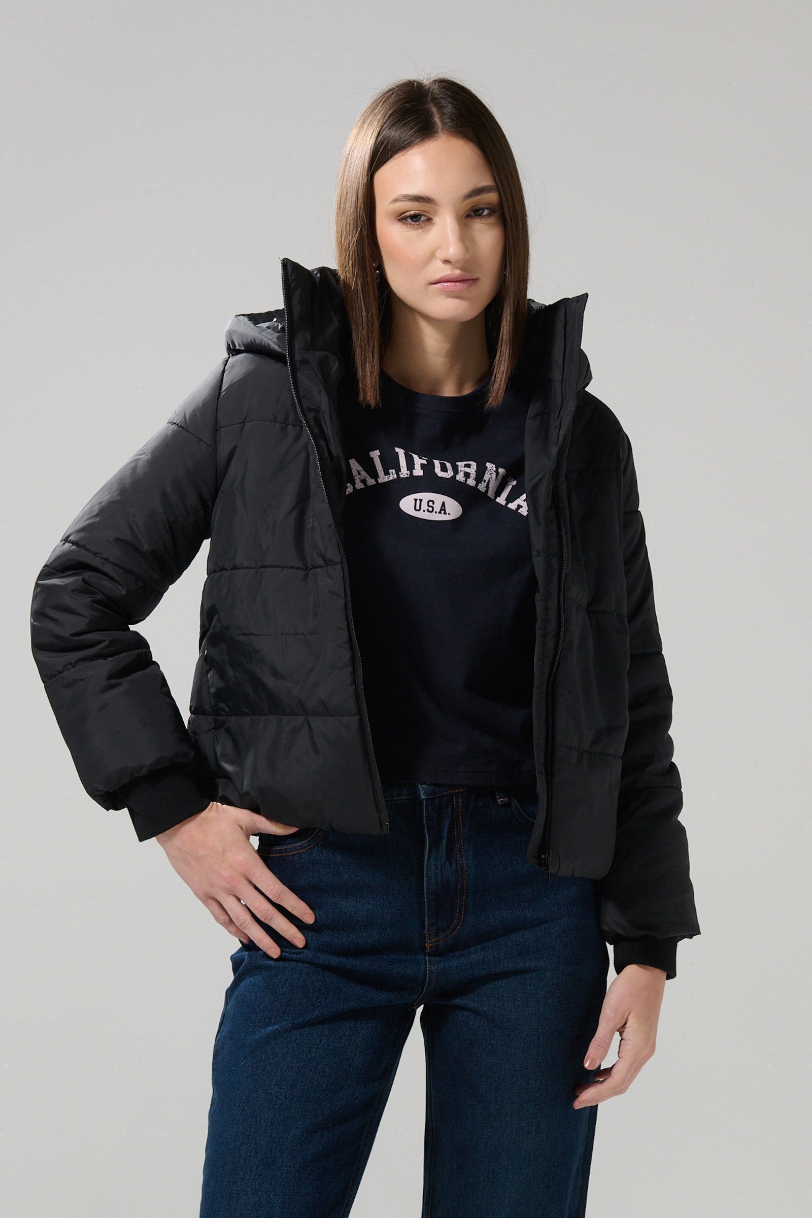 A woman with straight brown hair stands against a plain light background, looking at the camera with one hand on her hip. She is wearing a KOAJ CHAQUETA KOAJ CHARLOTTE 1 1/25, a black puffer jacket from KOAJ, layered over a navy California USA sweatshirt and blue jeans.