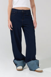 A person wearing the KOAJ PANTALON KOAJ JEAN STRAIGHT ROLL UP 3024—a dark blue, wide-leg jean from KOAJ—with extra denim fabric creating a stylish pooling effect around their feet, paired with white slip-on shoes and a white sleeveless crop top for a chic tiro bajo mujer ensemble.