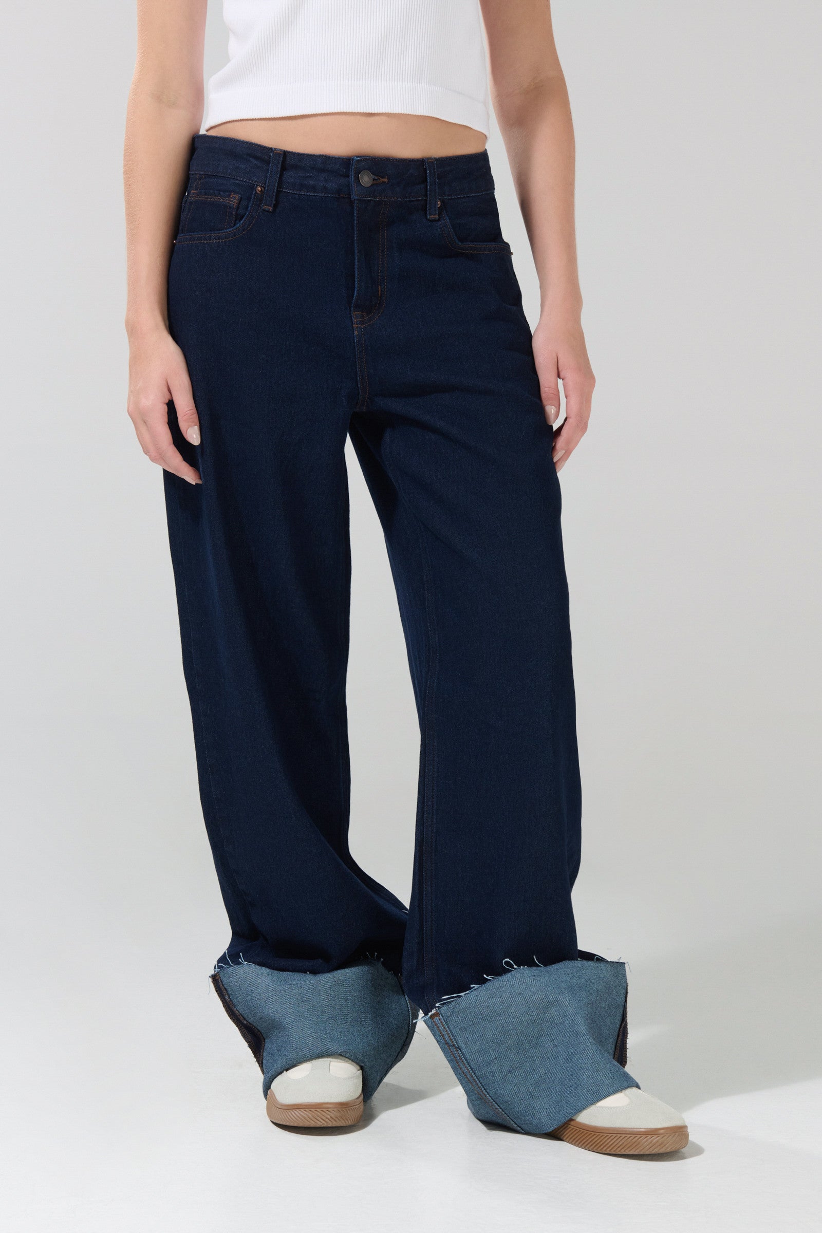 Wearing the KOAJ PANTALON KOAJ JEAN STRAIGHT ROLL UP 3024 by KOAJ, a person stands against a plain background. The straight, low-waist jeans in jean azul medio feature wide legs with large rolled denim cuffs that cover their feet and are paired with white slippers and a white crop top, adding a relaxed yet stylish touch to the overall look.