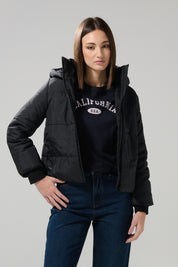 A woman wearing the KOAJ CHAQUETA KOAJ CHARLOTTE 1 1/25—a black puffer jacket from KOAJ—paired with dark jeans and a black California U.S.A. sweatshirt poses against a plain light gray background.