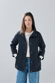 A woman stands facing forward against a plain light gray background, wearing the KOAJ CHAQUETA KOAJ CHARAMONYD 1 4/25 in navy by KOAJ over a white sweater and blue leopard-print jeans.