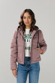 A woman with wavy brown hair is wearing a mauve KOAJ CHAQUETA KOAJ DASHY 1/25 by KOAJ, a white Michigan Varsity League sweatshirt, and blue jeans, standing against a plain light gray background.