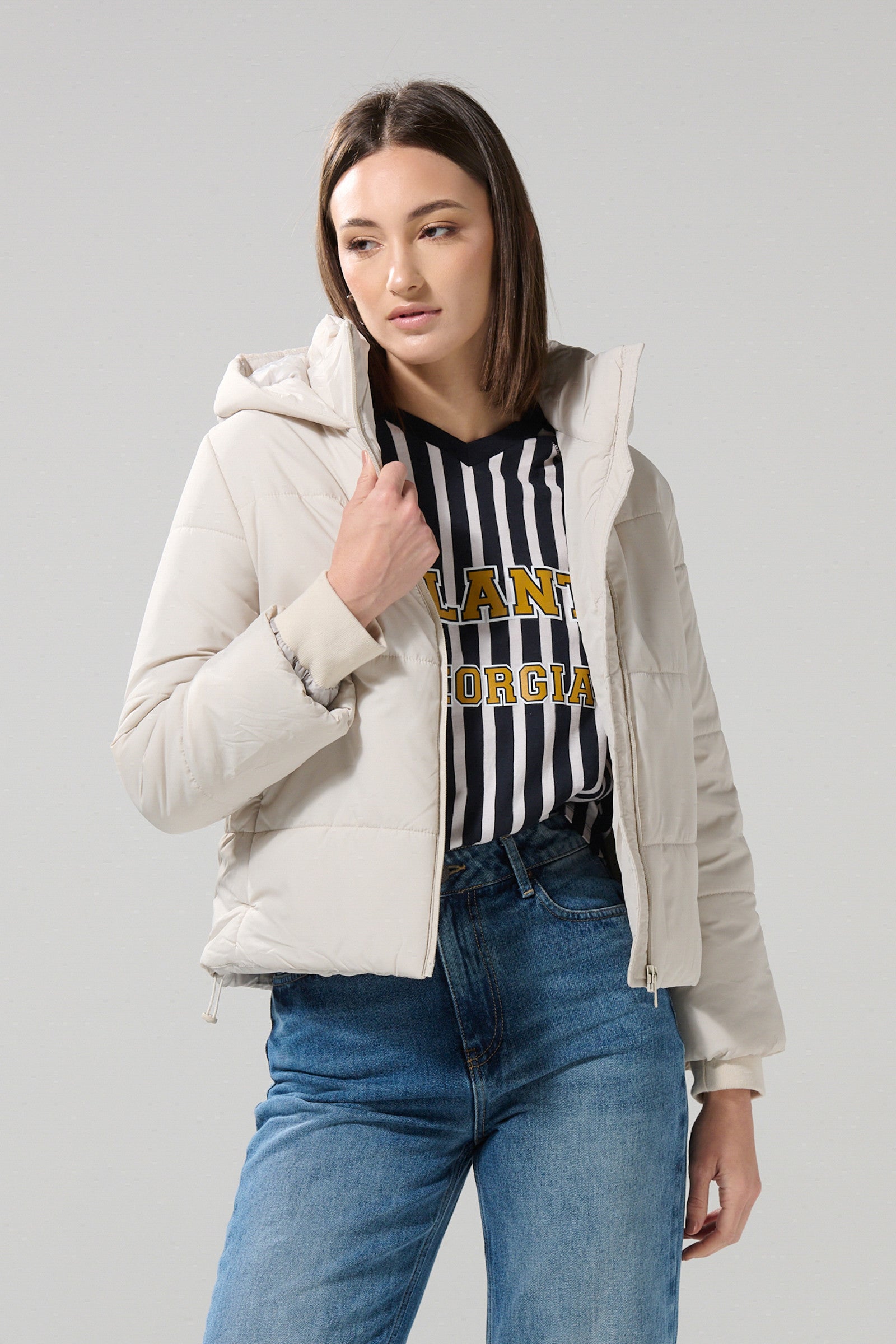 A woman wearing the KOAJ CHAQUETA KOAJ CHARLOTTE 1 1/25 by KOAJ over a black and white striped shirt and blue jeans stands against a plain background, looking to the side while holding the collar of her jacket.