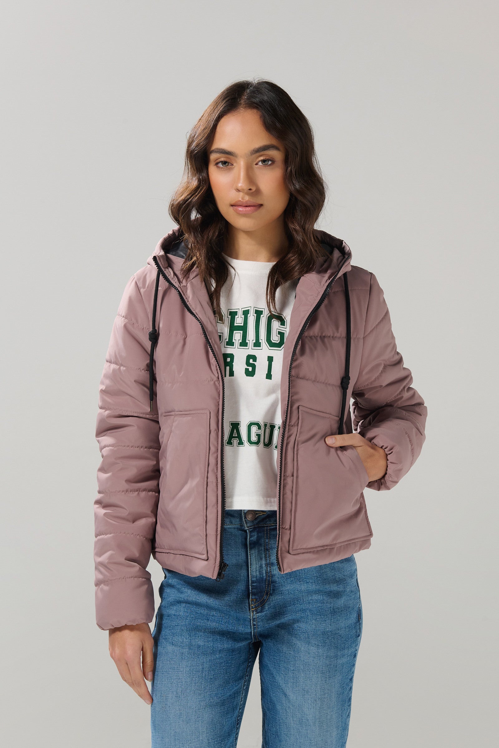 A woman wearing the KOAJ CHAQUETA KOAJ DASHY 1/25, a mauve quilted jacket from KOAJ, along with a white Michigan sweatshirt and blue jeans, stands against a light gray background, looking at the camera with a neutral expression.