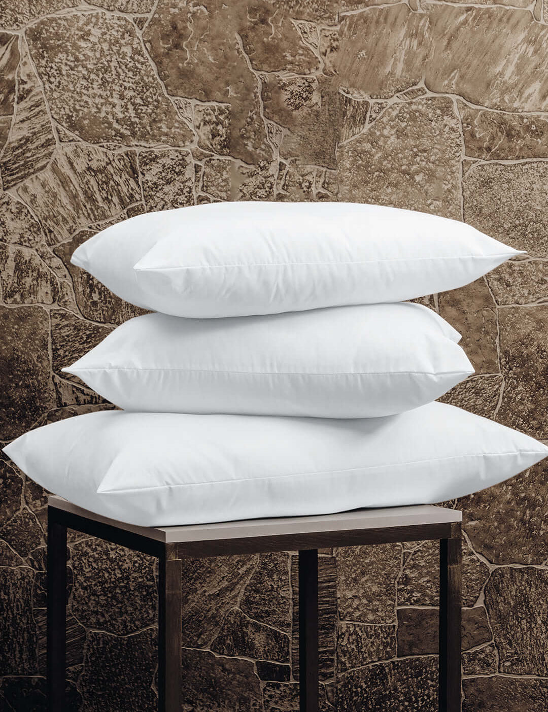 Three Almohada Microsuave by Intima Hogar, with soporte medio and in white, are stacked on a small dark wooden table, providing comfort against a stone-patterned brown wall.