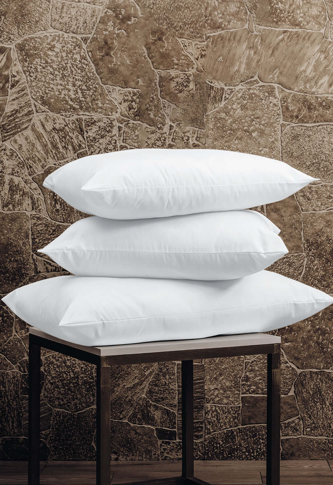Three Almohada Microsuave by Intima Hogar, with soporte medio and in white, are stacked on a small dark wooden table, providing comfort against a stone-patterned brown wall.