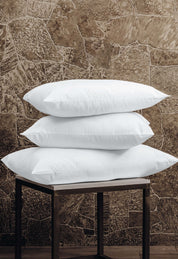 Three Almohada Microsuave by Intima Hogar, with soporte medio and in white, are stacked on a small dark wooden table, providing comfort against a stone-patterned brown wall.