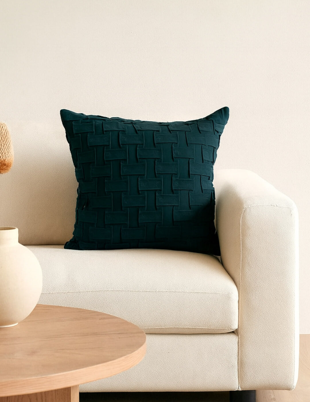 A Cojín de Lujo Brick Azul Petróleo de Intima Hogar con textura se luce en un moderno sofá color crema junto a una mesa redonda de madera clara y un florero beige, realzando la decoración elegante frente a una pared clara.