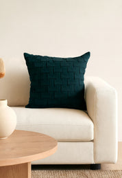 A Cojín de Lujo Brick Azul Petróleo de Intima Hogar con textura se luce en un moderno sofá color crema junto a una mesa redonda de madera clara y un florero beige, realzando la decoración elegante frente a una pared clara.