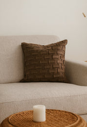 Un sofá beige decorado con cojines Brick Cafe de Intima Hogar en microfibra y relleno sintético. Frente al sofá, una mesa redonda de mimbre sostiene una vela blanca. Al fondo, la pared es lisa y clara.