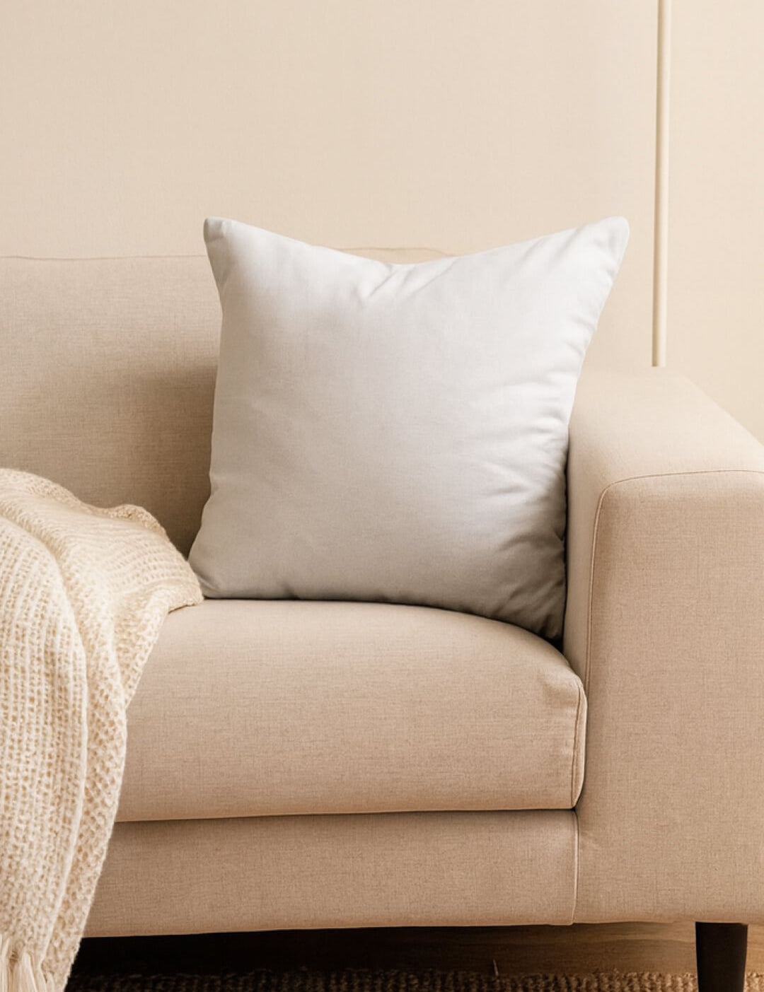 A light beige sofa features an Intima Hogar Cojín Cuadrado Blanco in the corner for extra soporte, with a cream textured blanket on the armrest. The neutral wall and carpet create a relaxing ambiente ideal for descanso.