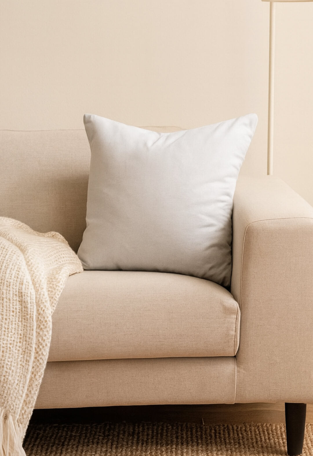 A light beige sofa features an Intima Hogar Cojín Cuadrado Blanco in the corner for extra soporte, with a cream textured blanket on the armrest. The neutral wall and carpet create a relaxing ambiente ideal for descanso.