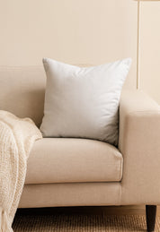 A light beige sofa features an Intima Hogar Cojín Cuadrado Blanco in the corner for extra soporte, with a cream textured blanket on the armrest. The neutral wall and carpet create a relaxing ambiente ideal for descanso.