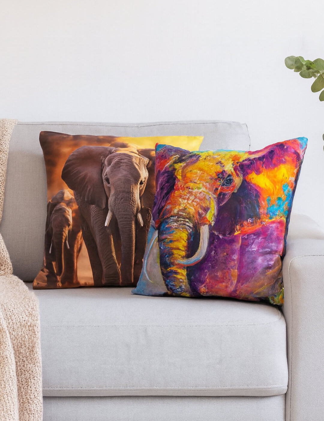Dos cojines de Intima Hogar adornan el sofá: uno es el "Cojín Elefante" con elefantes al atardecer y el otro luce un diseño colorido. Se aprecia además una manta beige y una planta verde.