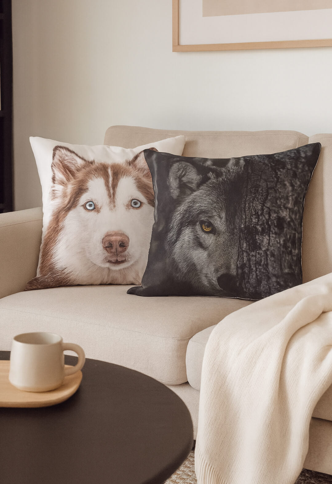 On a beige couch, two cojines decorativos—including Intima Hogar’s Cojín Estampado Lobo—add visual flair. A cream throw and a round coffee table with a cup complete the cozy setting.