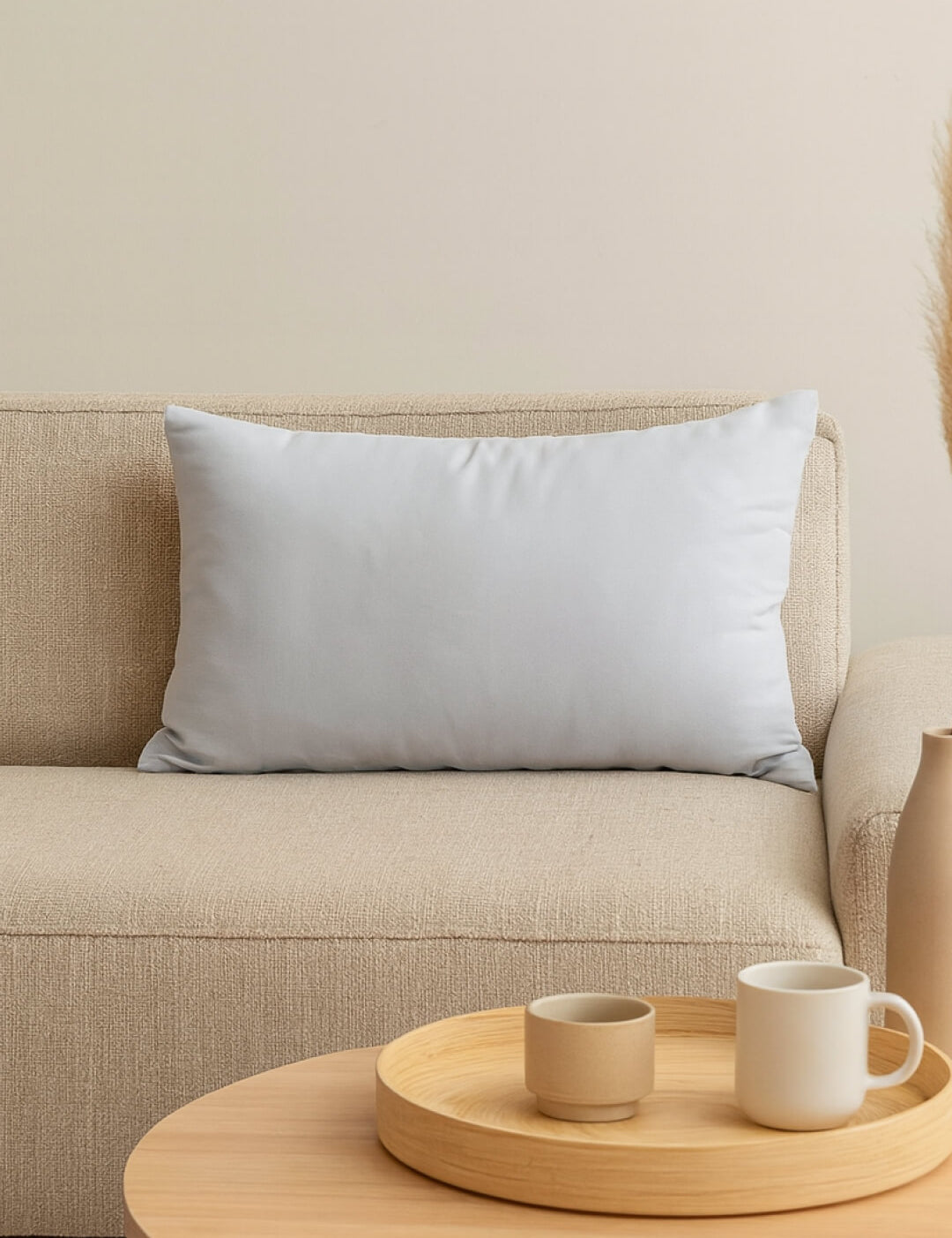 A Cojín Rectangular Blanco de Intima Hogar descansa sobre un sofá beige, brindando soporte y confort. Delante, una mesa de centro redonda sostiene una bandeja con taza, cuenco y mug. El ambiente es minimalista y acogedor.