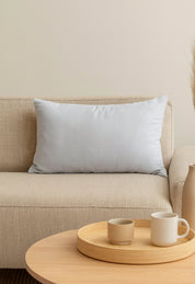 A Cojín Rectangular Blanco de Intima Hogar descansa sobre un sofá beige, brindando soporte y confort. Delante, una mesa de centro redonda sostiene una bandeja con taza, cuenco y mug. El ambiente es minimalista y acogedor.