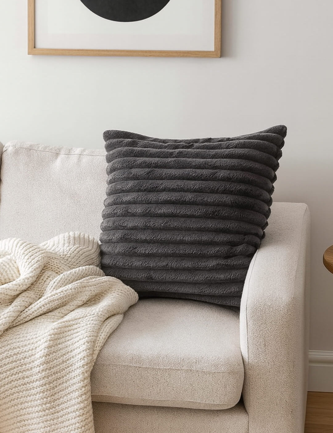 The Cojín Sultan by Intima Hogar in textured dark gray rests on a light beige sofa next to a cream knit blanket, while a framed abstract art print above enhances the modern style.