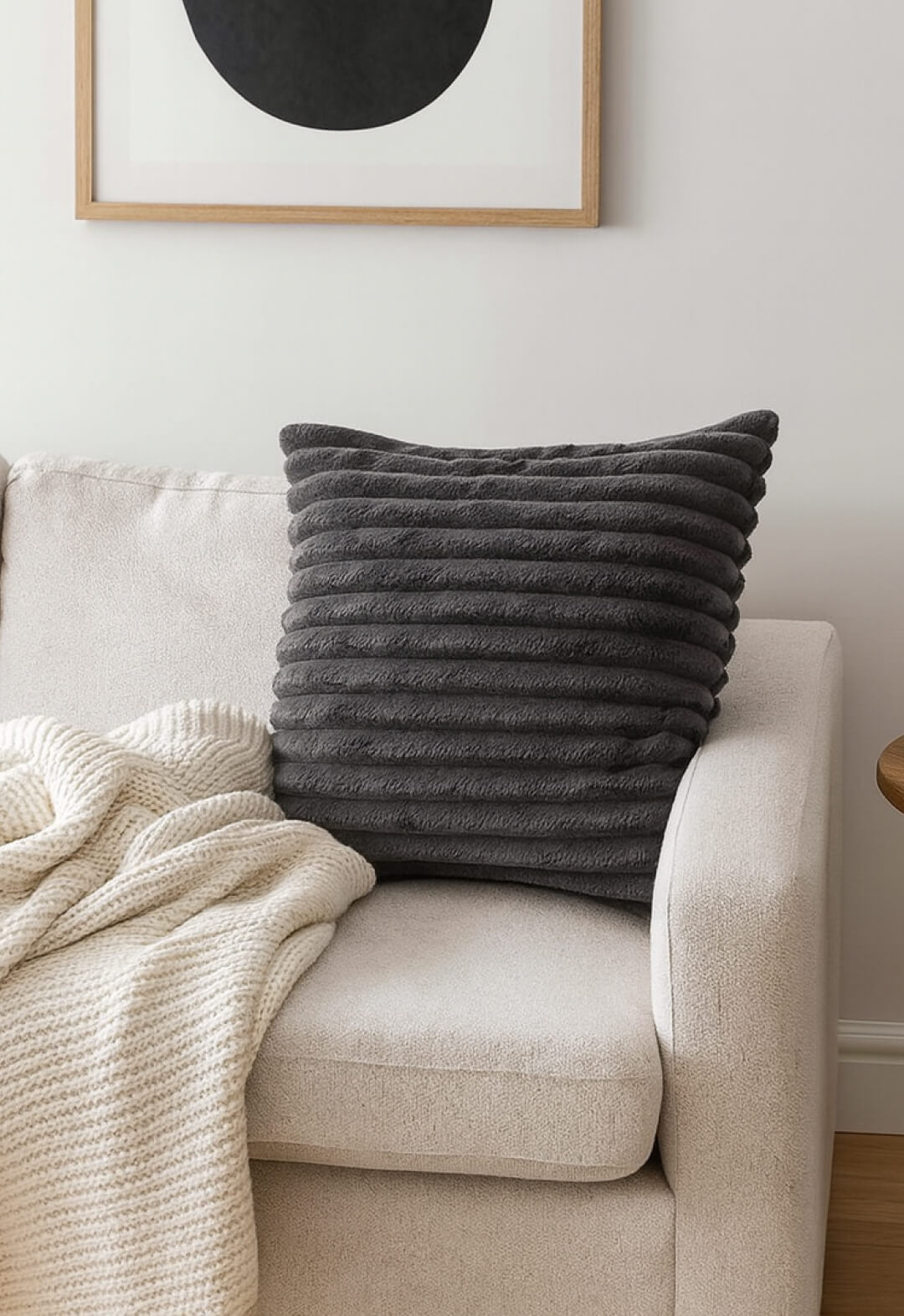 The Cojín Sultan by Intima Hogar in textured dark gray rests on a light beige sofa next to a cream knit blanket, while a framed abstract art print above enhances the modern style.