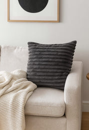 The Cojín Sultan by Intima Hogar in textured dark gray rests on a light beige sofa next to a cream knit blanket, while a framed abstract art print above enhances the modern style.
