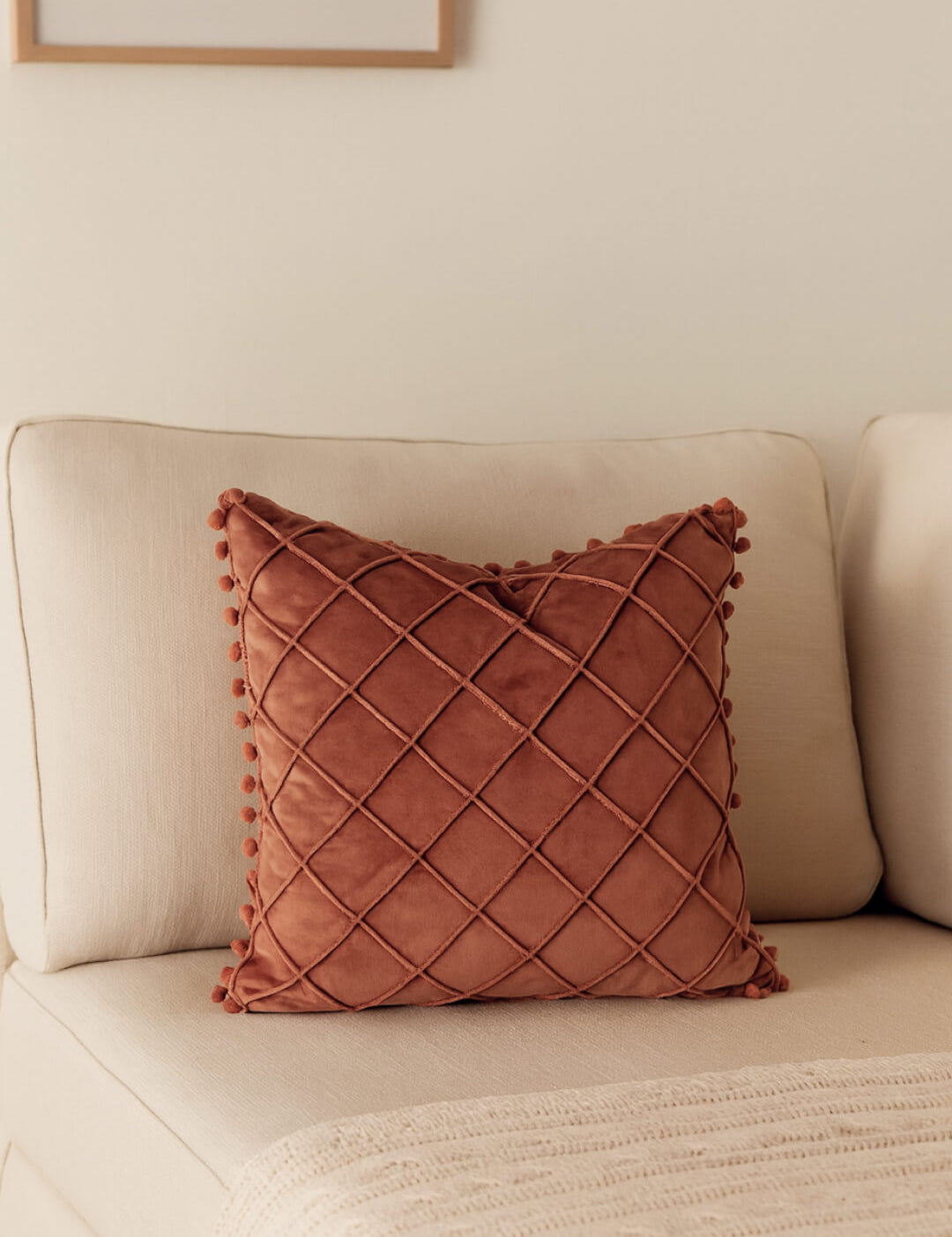 The Cojín de Lujo Velvet Rosa by Intima Hogar, featuring a diamond quilted pattern and pom-pom edges, rests on a cream sofa beside a beige knit blanket in a softly lit, neutral-toned living room.