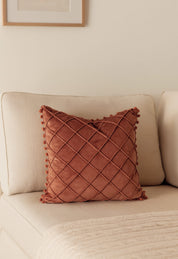 The Cojín de Lujo Velvet Rosa by Intima Hogar, featuring a diamond quilted pattern and pom-pom edges, rests on a cream sofa beside a beige knit blanket in a softly lit, neutral-toned living room.