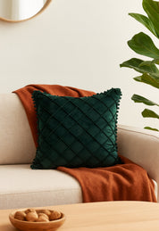 El Cojín de Lujo Velvet Verde Esmeralda de Intima Hogar reposa sobre un sofá marfil con manta naranja quemada; en la mesa, bol de madera con nueces, y al fondo una planta verde y un espejo redondo decoran el ambiente.