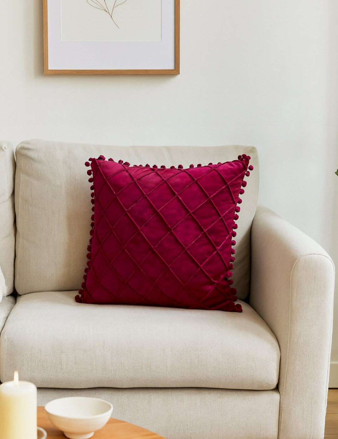 A beige sofa with the Intima Hogar Cojín de Lujo Velvet Vino, featuring a rich quilted design and pom-pom edges, sits by a wooden coffee table with a candle and bowl, plus a minimalist art print—añadiendo un toque sutil de lujo.