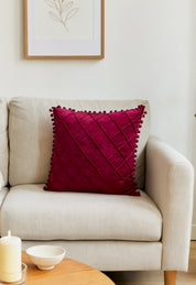A beige sofa with the Intima Hogar Cojín de Lujo Velvet Vino, featuring a rich quilted design and pom-pom edges, sits by a wooden coffee table with a candle and bowl, plus a minimalist art print—añadiendo un toque sutil de lujo.