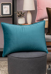 A rectangular Funda de Almohada Ensueño Verde by Intima Hogar rests on a round gray ottoman. Behind it, decor accents fill a shelf, with a white lamp and a pink pillow on the bed, creating a cozy, modern bedroom ambiance.