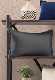 La Funda de Cojín Rectangular Sueños Gris Oxford de Intima Hogar, con textura suave, descansa sobre una repisa oscura junto a un jarrón decorativo blanco con patrones negros y un cojín azul; abajo se ve una planta y otro cojín verde claro.