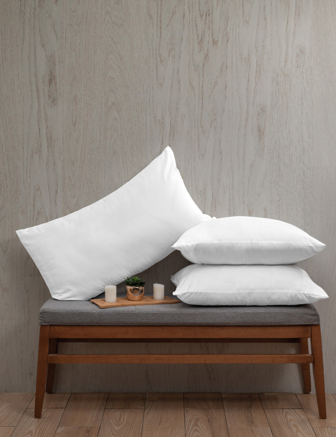 Three white Almohada Microsuave pillows by Intima Hogar with medium support are stacked on a gray cushioned bench with wooden legs. Small candles and a potted plant in front add comfort to the light wood floor and textured wall backdrop.