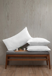 Three white Almohada Microsuave pillows by Intima Hogar with medium support are stacked on a gray cushioned bench with wooden legs. Small candles and a potted plant in front add comfort to the light wood floor and textured wall backdrop.