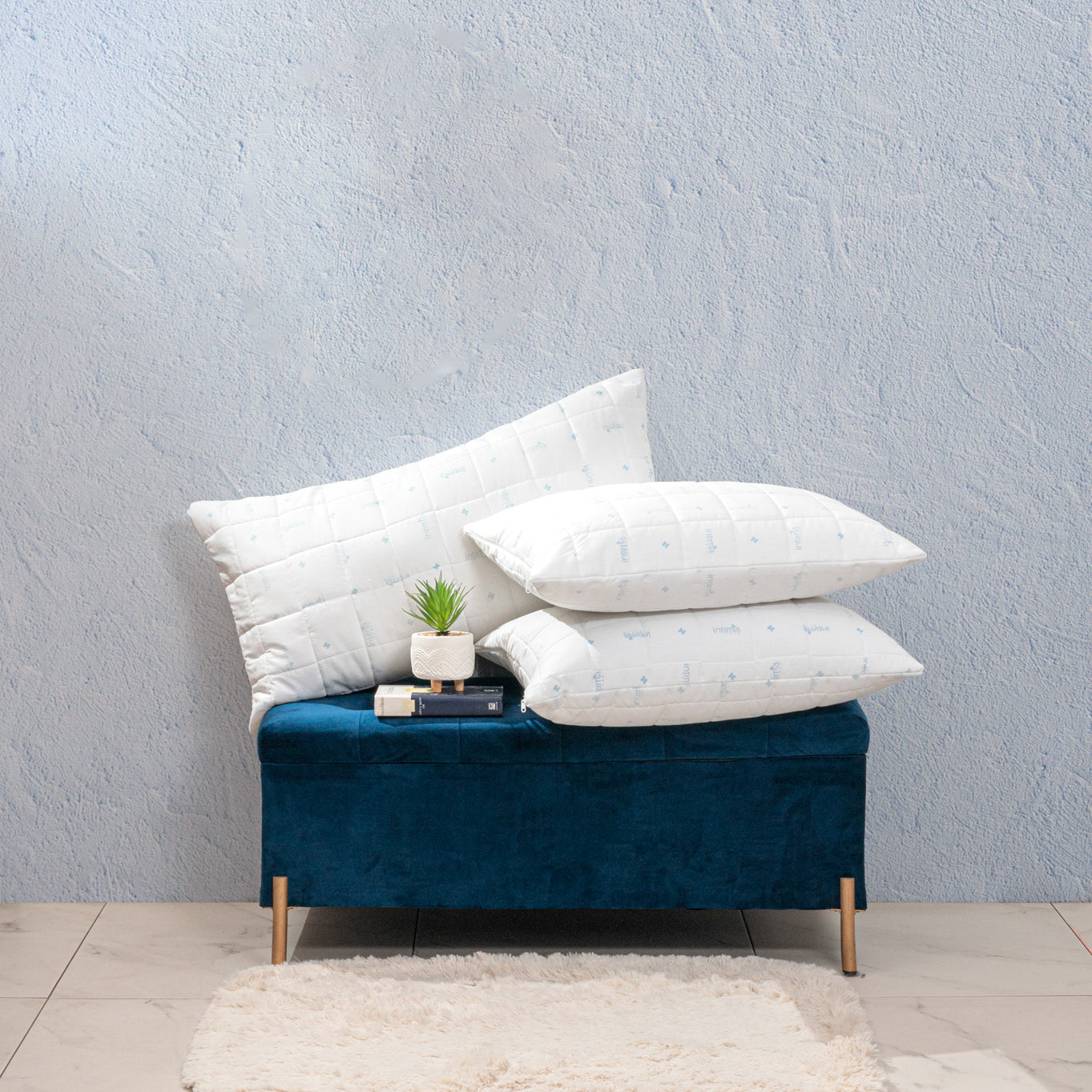 A blue upholstered bench with three Intima Hogar Almohada Protección Plus pillows stacked on it, a small potted plant and books on top; a white rug lies on the tiled floor beneath, set against a textured light blue wall.