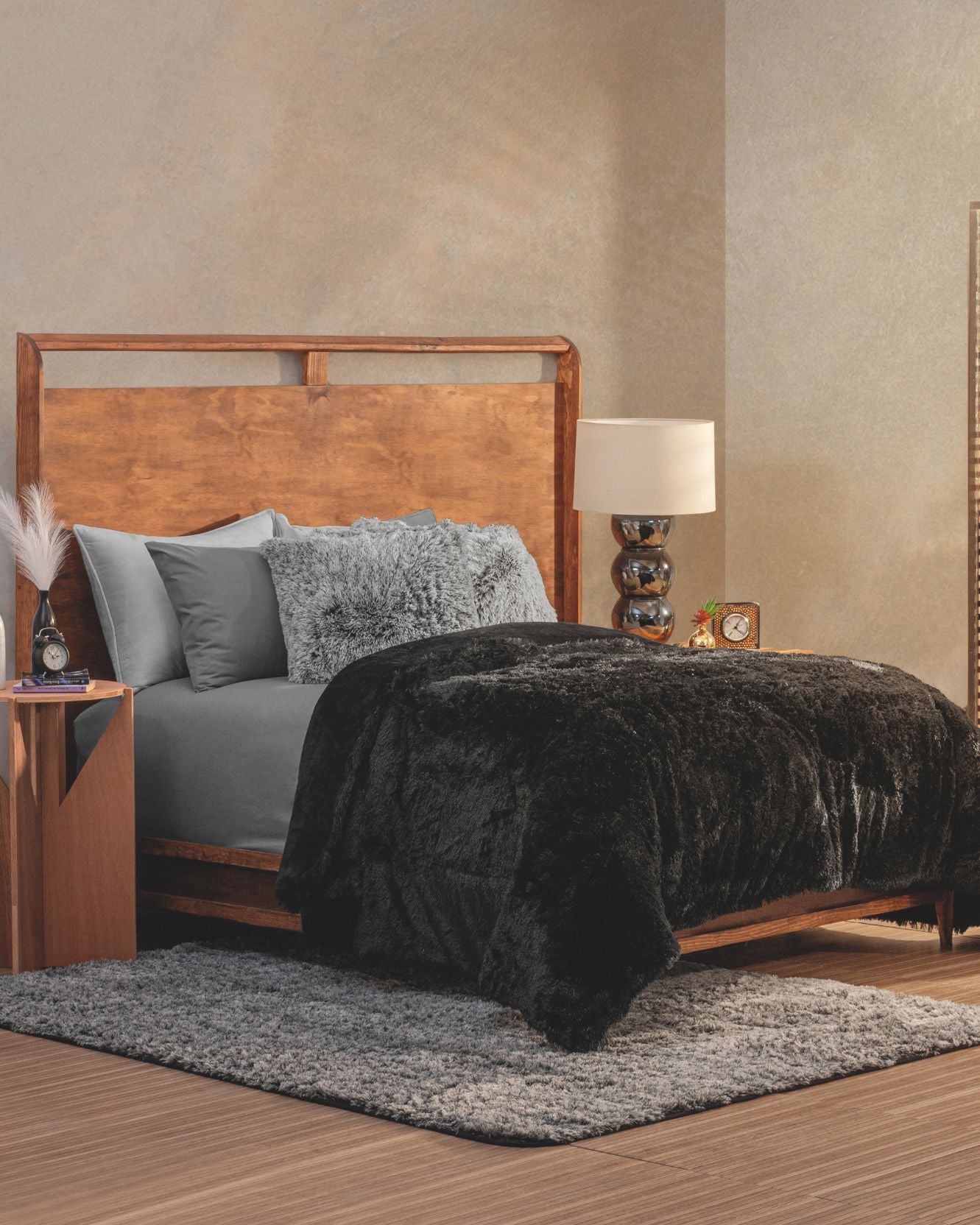 A modern, elegant habitación with a wooden bed frame, gray bedding, fluffy pillows, and a Cobertor Platinum Extra Suave Albania by Intima Hogar in black atop the bed, plus a wooden nightstand, lamp, and textured gray rug over wood flooring.