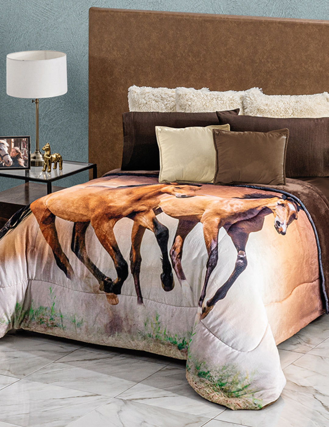 A neatly made bed with a brown headboard, white and brown pillows, and the Cobertor Digital Ultra Suave Caballos by Intima Hogar featuring running horses. The room includes a nightstand with a lamp, decorative horse, and photo frame.