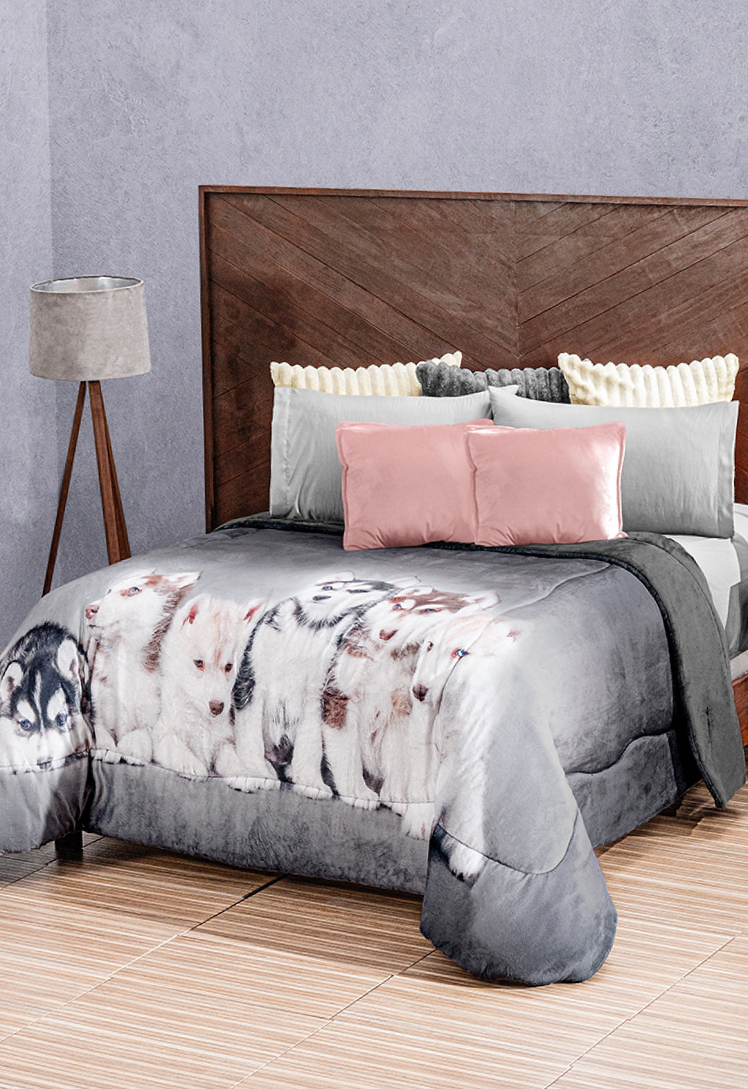 A neatly made bed features the Intima Hogar Cobertor Digital Ultra Suave Huskies and cozy gray, white, and pink pillows. A wooden headboard and floor lamp complement the look against a textured gray wall.