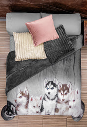 A bed with gray sheets, a pink pillow, textured beige and gray pillows, and the Cobertor Digital Ultra Suave Huskies by Intima Hogar featuring five huskies. Viewed from above next to a wooden floor and dark nightstand.