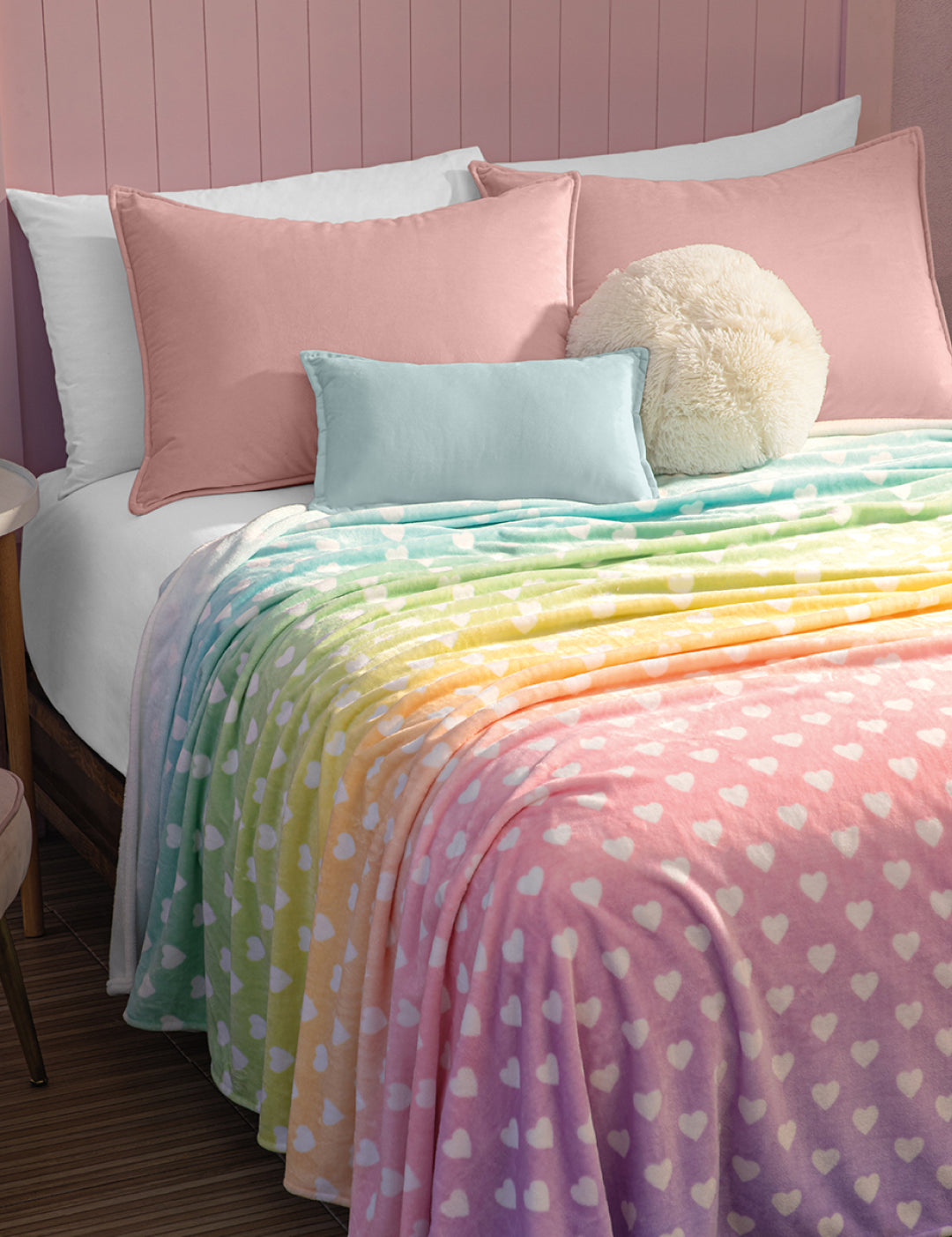 A neatly made bed with pink pillows, a light blue pillow, and a round cream cushion is topped with the Intima Hogar Cobertor Flannel Ligero Azúcar, featuring a pastel rainbow heart design, set against a charming pink paneled wall.