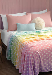A neatly made bed with pink pillows, a light blue pillow, and a round cream cushion is topped with the Intima Hogar Cobertor Flannel Ligero Azúcar, featuring a pastel rainbow heart design, set against a charming pink paneled wall.