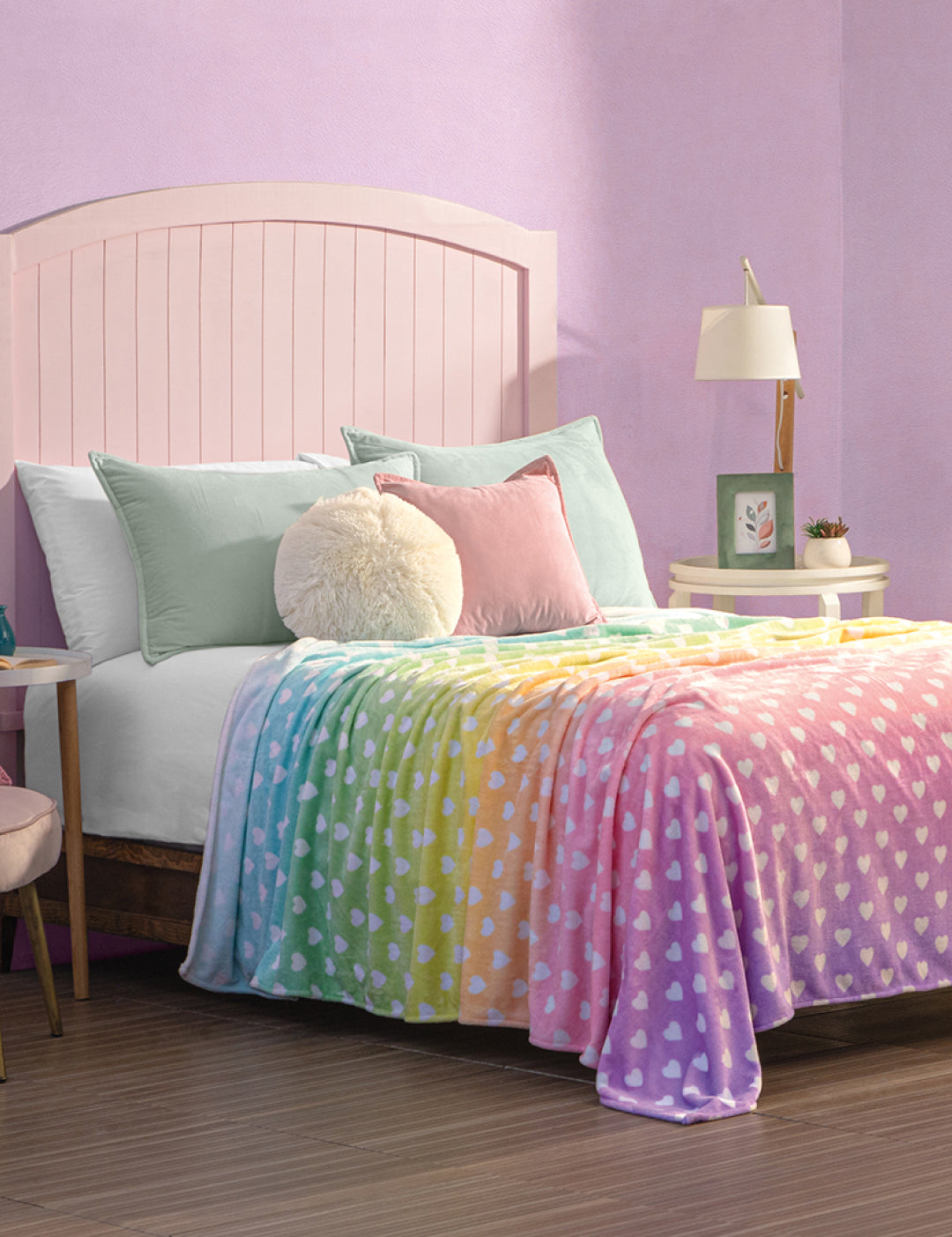 A cozy bedroom features a light pink headboard bed, pastel pillows, and the Cobertor Flannel Ligero Azúcar by Intima Hogar—a rainbow-colored blanket with white heart designs. Purple walls and nightstands with lamps, plants, and decor add extra charm.
