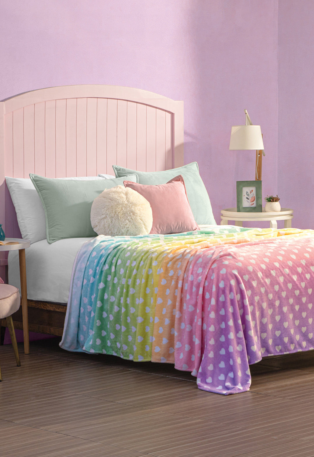 A cozy bedroom features a light pink headboard bed, pastel pillows, and the Cobertor Flannel Ligero Azúcar by Intima Hogar—a rainbow-colored blanket with white heart designs. Purple walls and nightstands with lamps, plants, and decor add extra charm.